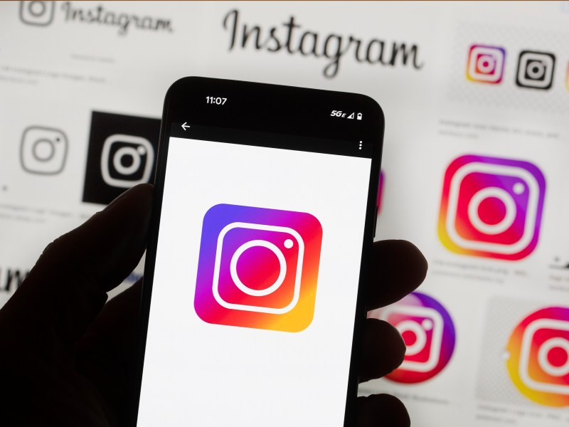 A hand holds a smartphone displaying the Instagram logo, with various versions of the Instagram logo shown on a screen in the background.