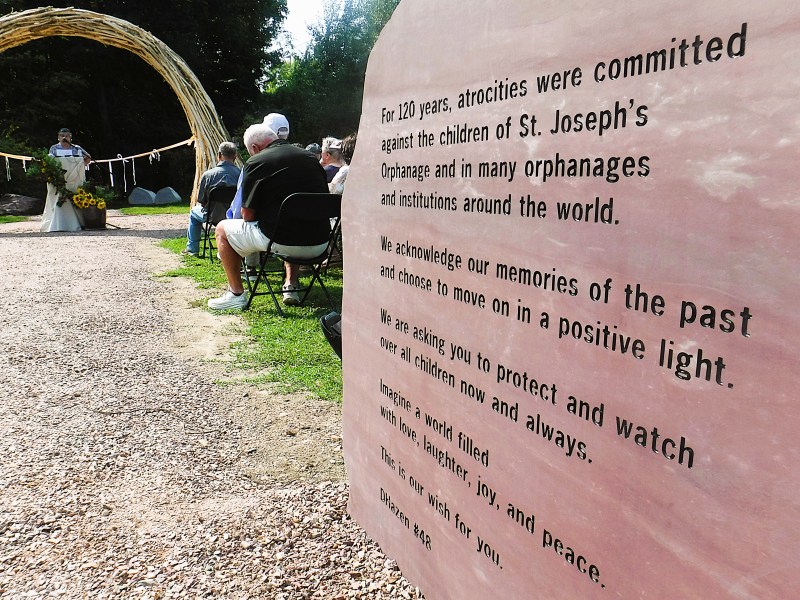 A memorial event is held outdoors near a large plaque acknowledging atrocities against children at St. Joseph’s Orphanage and other institutions, urging protection and remembrance.