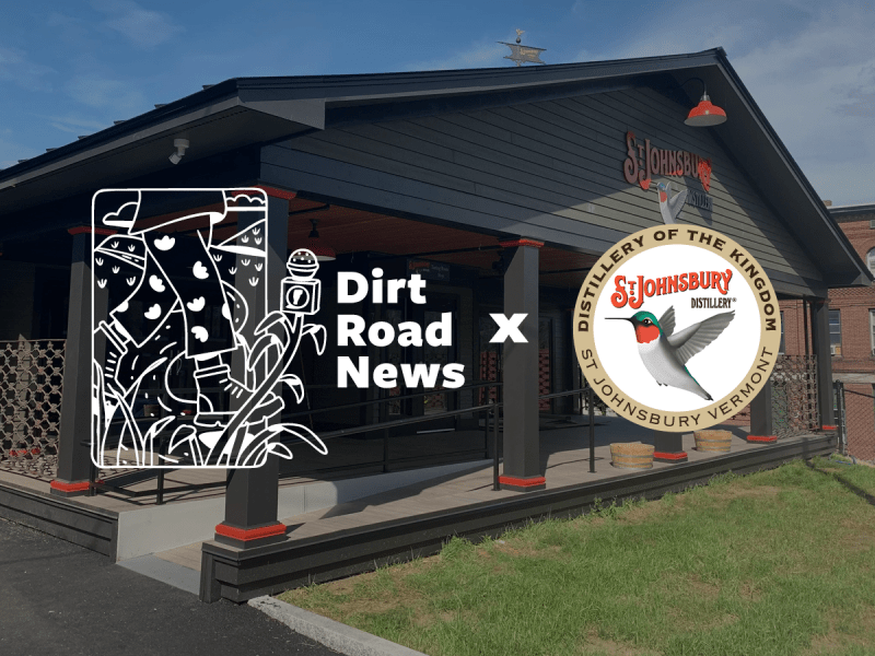 A black building with a porch displays two logos: Dirt Road News and St. Johnsbury Distillery, indicating a collaboration.