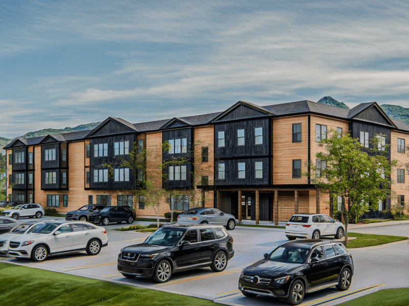 A three-story apartment building with wood and dark paneling, surrounded by parked cars and greenery, with mountains in the background under a cloudy sky.